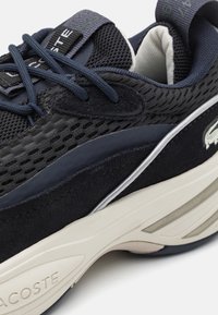 Gros plan d'une basket Lacoste noire et bleu marine avec tige en mesh et daim, lacets foncés, semelle intermédiaire blanche amortie et logo sur le talon et la languette.