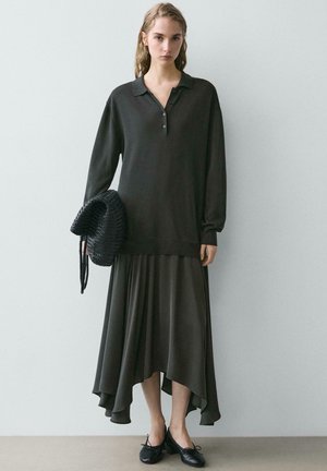 Młoda kobieta ubrana w czarną koszulkę polo z długim rękawem i asymetryczną czarną spódnicę, trzymająca plecioną czarną torbę, stojąca na tle jasnej ściany.