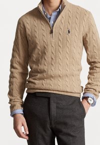 Homme portant un pull beige en maille torsadée à demi-fermeture éclair sur une chemise rayée bleue, avec un pantalon gris charbon, une montre au poignet gauche, la main droite dans la poche.