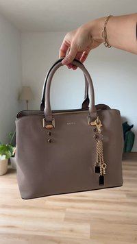 Grå syntetisk håndveske med gullhardware, doble håndtak, glidelås-lukking og dekorativt nøkkelringsanheng med charms. Glatt tekstur og strukturert design.