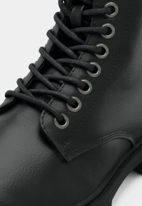 Bottine en cuir noir avec lacets ronds, œillets métalliques, texture lisse et semelle discrète. Couture détaillée sur les côtés.