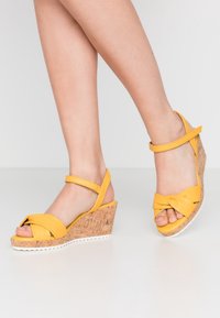 Gula kilklackar med korkklack, med en knutdesign på remmarna och justerbar ankelrem. Slät yta med vit sula.