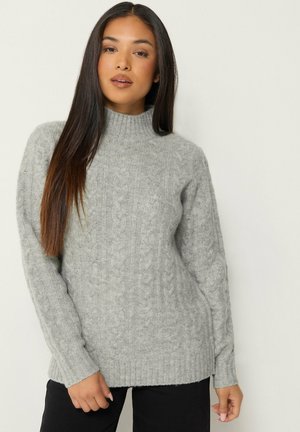 Femme aux longs cheveux bruns et raides portant un pull à col roulé gris clair à torsades et un pantalon noir sur un fond uni.
