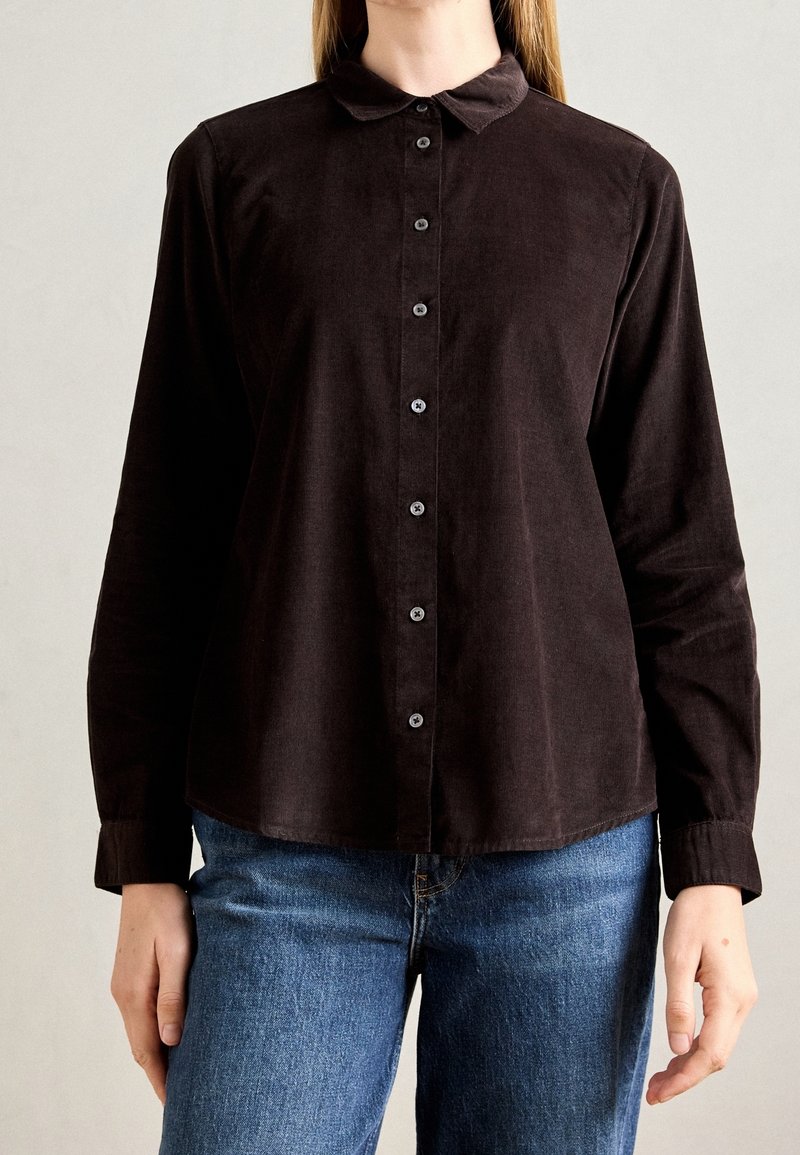 Personne portant une chemise noire à manches longues avec col et un jean bleu sur un fond clair uni.