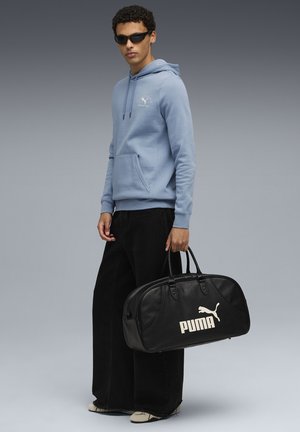 Lyseblå hoodie med frontlomme, sorte brede bukser og en sort læder rejsetaske med et hvidt Puma-logo. Solbriller bæres.