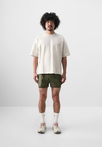 Beige oversized t-shirt med rund hals och subtila tryck, kombinerad med olivgröna shorts och vita strumpor. Skorna är ljusa sportskor.