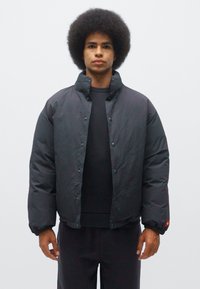 Black padded jacket with a high collar, snap button closure, elastic cuffs, and a smooth texture. Features a small red logo on the sleeve.