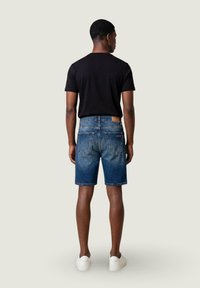Hombre de pie de espaldas, vistiendo camiseta negra, pantalones cortos de mezclilla azul y zapatillas blancas sobre un fondo claro y liso.