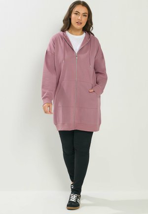 Frau trägt einen langen mauvefarbenen Kapuzenpullover mit Reißverschluss, weißes Hemd, schwarze Leggings und schwarze Turnschuhe, geht vor einem schlichten hellen Hintergrund.