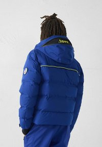 Person mit Dreadlocks, die eine blaue Steppjacke mit Kapuze und einem Logoaufnäher am linken Ärmel trägt, steht vor einer schlichten hellen Wand.