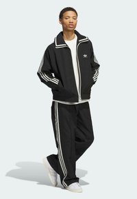 Schwarze Sportjacke und -hose mit weißen Streifen, mit Reißverschluss und Fronttaschen. Wird über einem weißen T-Shirt getragen und mit weißen Turnschuhen kombiniert.