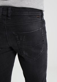 Acercamiento de una persona que lleva puestos unos jeans negros desgastados con detalles desgastados en el bolsillo trasero y una camisa gris claro metida por dentro.