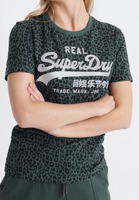 Ciemnozielona koszulka z czarnym wzorem lamparta, z wyraźnym białym logo "SuperDry" oraz japońskim tekstem na środku.