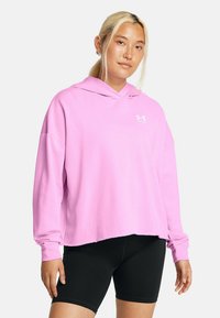 Sudadera rosa con corte holgado y mangas largas, con un bolsillo canguro y detalle del logo en blanco en el pecho. Combinada con pantalones cortos negros.