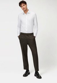 Witte overhemd met knopen en kraag, gecombineerd met donkergroene broeken met elastische taille en zwarte schoenen. Eenvoudig, strak design.