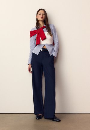 Kvinde iført en blåstribet skjorte med en hvid korsettop over og en rød sweater bundet om skuldrene, marineblå bukser og sorte sko.