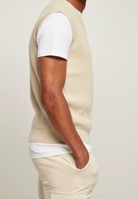 Beżowy, dziany sweter bez rękawów o prążkowanej teksturze, noszony na białej koszulce. Model ma ręce w kieszeniach jasnobeżowych spodni.