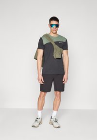T-shirt nera a maniche corte con dettagli verdi, abbinata a pantaloni corti neri. Il modello indossa occhiali da sole blu e sneaker chiare.