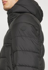 Sort dunjakke med hætte, med quiltet design og blød, glat tekstur. Inkluderer en cirkulær logo på skulderen og sidelomme.