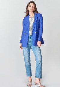 Blazer de lino azul con un diseño de doble botonadura y botones dorados, combinado con una blusa a rayas y jeans claros desgastados.