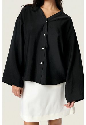 Vrouw draagt een losse zwarte blouse met knopen en brede mouwen, gecombineerd met een witte rok tot op de knie, tegen een effen achtergrond.