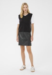 Gilet in maglia nera con chiusura a bottoni sopra una t-shirt bianca, abbinato a una gonna in pelle nera e sneakers crema con dettagli neri.