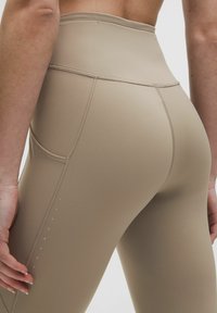 Beige színű leggings, magas derékkal, sima textúrával, oldalsó zsebbel és kontrasztos varrással. Az külső varrat mentén apró pöttyökkel díszített.
