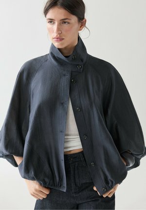 Frau trägt eine dunkle, strukturierte, geknöpfte Jacke mit hohem Kragen und Ballonärmeln, kombiniert mit dunkler Hose und einem hellen, kurz geschnittenen Oberteil.