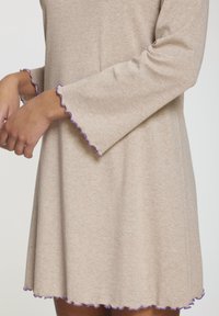 Vestido de color beige claro con mangas largas, que presenta bordes morados ondulados en los puños y el dobladillo. Fabricado con una suave tela texturizada.