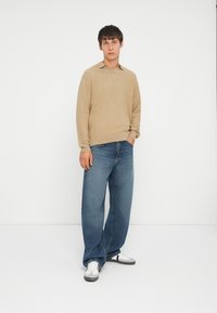 Beżowy sweter z dzianiny z kołnierzykiem, w zestawieniu z luźnymi niebieskimi dżinsami. Model nosi białe trampki z szarymi akcentami na jednolitym tle.