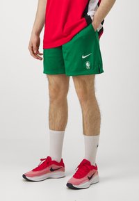 Rohelised Nike korvpallilühikesed püksid, millel on võrkkangas ja küljetaskud, koos punaste Nike tossudega, valgete sokkidega ja punase kampsuniga.