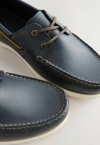 Námořnicky modré kožené lodní boty s bílým prošitím, hnědými koženými tkaničkami a bílými gumovými podrážkami na neutrálním pozadí.