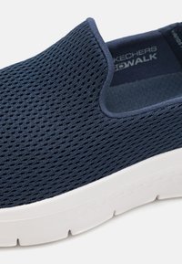 Scarpa slip-on in rete blu navy con suola bianca liscia, colletto elasticizzato e discreto branding testuale all'interno.