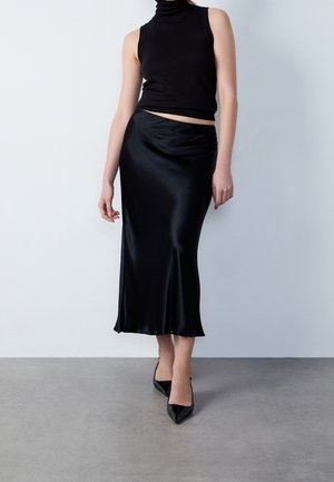 Frau trägt ein ärmelloses schwarzes Rollkragenoberteil, einen schwarzen Satin-Midirock und schwarze spitze High Heels vor einem schlichten hellen Hintergrund.