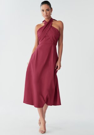 Burgundy-holmaloha mekko, jossa on kierretty yläosa, soljuva A-linjainen hame ja pehmeä tekstuuri. Minimalistinen muotoilu ilman näkyviä kuvioita tai koristeita.