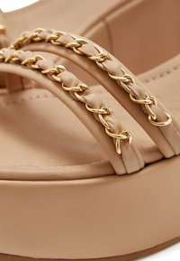 Beige platform sandalen met gevlochten leren riemen, voorzien van gouden kettingaccenten, een gladde textuur en een gevormde zool.