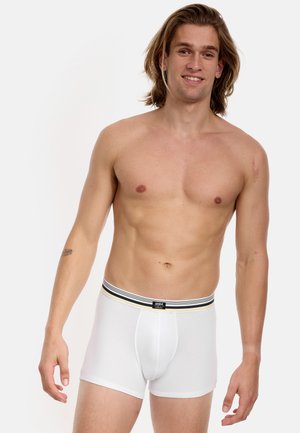 Weißer Boxerslip mit einem schwarz-gelben gestreiften Bund, der eng anliegt und eine glatte Textur aufweist. Getragen von einem oberkörperfreien männlichen Modell.
