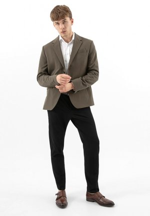 Młody mężczyzna poprawiający mankiety, ubrany w oliwkową marynarkę, białą koszulę, czarne spodnie i brązowe eleganckie buty, stojący na białym tle.