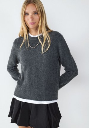 Jonge vrouw met lang blond haar die een donkergrijze trui, wit ondershirt, zwarte rok en een zilveren ketting draagt tegen een effen achtergrond.