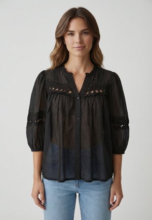 Giovane donna che indossa una camicetta nera trasparente con bottoni, dettagli in pizzo e maniche a tre quarti, abbinata a jeans azzurri, in piedi su uno sfondo bianco.