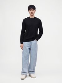 Jeune homme debout portant un pull noir à manches longues, un jean large bleu clair et des baskets blanches sur un fond blanc uni.