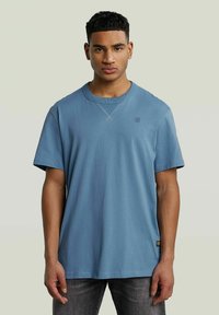 Niebieska bawełniana koszulka z krótkim rękawem, okrągłym dekoltem i subtelnym detalem logo na piersi. Materiał wydaje się być miękki i lekko teksturowany.