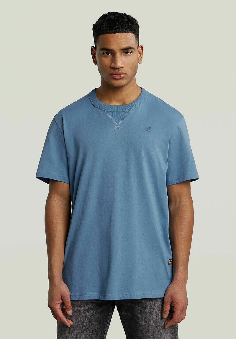 Niebieska bawełniana koszulka z krótkim rękawem, okrągłym dekoltem i subtelnym detalem logo na piersi. Materiał wydaje się być miękki i lekko teksturowany.