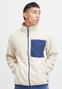 Fleecejacke in Creme mit einer marineblauen, mit Reißverschluss versehenen Brusttasche. Verfügt über einen hohen Kragen und einen durchgehenden Frontreißverschluss. Weiche Textur, legere Passform.