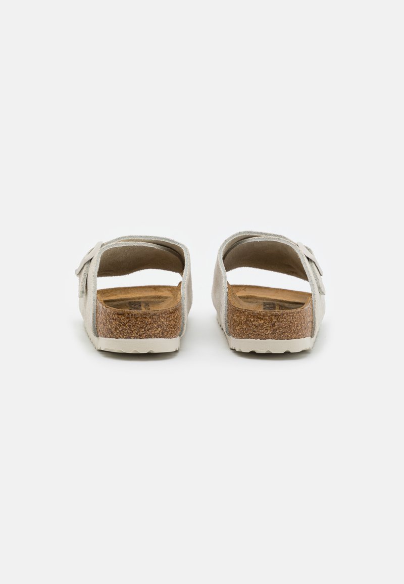 Hellgraue Wildleder-Slipper mit Korkfußbett und weißer Sohle, inklusive verstellbarem Riemen mit Schnalle. Rückansicht dargestellt.
