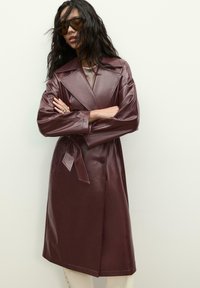Bordová trench coat s dlhým rukávom a opaskom v páse, širokými klopami a lesklým umelým koženým materiálom. Obsahuje dve predné vrecká a gombíkový uzáver.