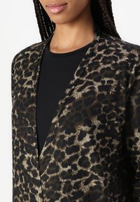 Leopardmönstrad kavaj i mjukt, lättviktsmaterial, med öppen framsida och en svart underliggande topp; har jordtoner och kontrasterande texturer.