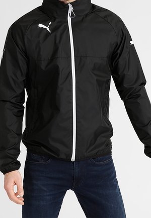 Veste coupe-vent noire Puma avec fermeture éclair blanche et logos sur la poitrine et les manches portée par une personne en jean bleu foncé.