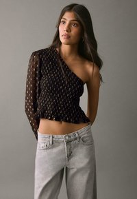 Top nero con scollo a barchetta, dettagli arricciati e manica trasparente con motivo; abbinato a jeans denim grigio chiaro, larghi in vita.