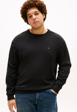 Jeune personne aux cheveux bouclés portant un t-shirt noir à manches longues et un jean bleu, debout avec une main dans la poche sur fond blanc.